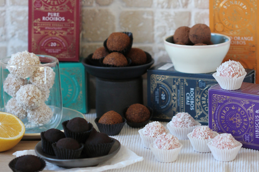 Selection of five types of chocolate truffles, each made with a different flavoured tea, beautifully displayed on a selection of tiny plates and crockery in front of the tea products that were used to make the truffles.